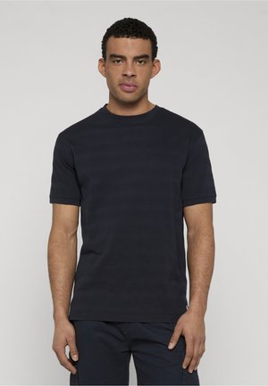 Homme debout, faisant face à l'avant, portant une chemise à manches courtes rayée bleu marine foncé et un pantalon assorti bleu marine, sur un fond clair uni.