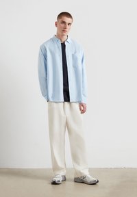 Camicia button-up azzurro chiaro con un singolo taschino sul petto, indossata sopra una maglietta scura, abbinata a pantaloni bianchi a gamba larga e sneakers argento.