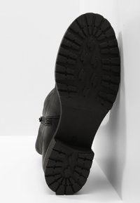 Semelle de botte noire avec un motif de bande de roulement épais reposant contre un mur d’angle blanc et un sol blanc.