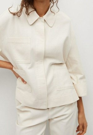 Osoba ubrana w luźną, kremowo-białą koszulę na guziki z dużymi kieszeniami oraz dopasowane spodnie na jednolitym tle.