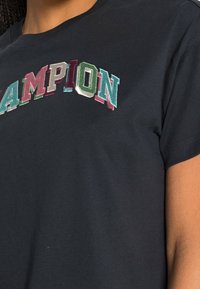 Svart bomullströja med flerfärgad "CHAMPION" broderi i fet stil på framsidan. Korta ärmar och rundad halsringning.