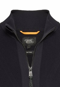 Sort strikket zip-up sweater med høj krave, der har et tekstureret mønster, en kontrastfarvet orange label og en metal lynlås.