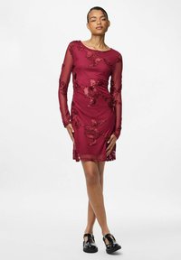 Žena v těsně přiléhajících bordó šatech s květinovou výšivkou, průsvitnými dlouhými rukávy a černými botami Mary Jane, stojící před bílým pozadím.