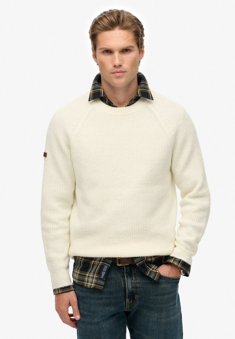 Cremiger gerippter Pullover mit Raglanärmeln, über einem grün-blauen Karohemd getragen. Kombiniert mit blauen Jeans und einem Ledergürtel.
