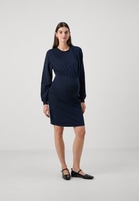 Rochie de maternitate bleu marin cu mâneci lungi, talie adunată și textură fină. Asortată cu balerini negri. Design simplu, ajustat.