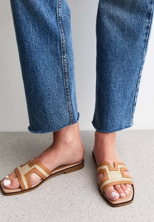 Sandali slide marroni e beige con un design geometrico, realizzati in materiale intrecciato, abbinati a jeans in denim blu con risvolti.