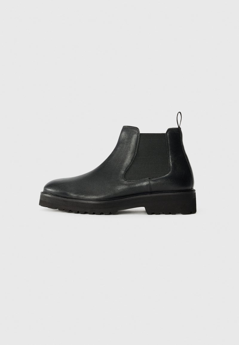 Botte Chelsea en cuir noir avec semelle épaisse, panneaux latéraux élastiques et languette à l'arrière, vue de profil sur fond gris.