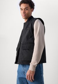 Schwarze ärmellose Jacke mit strukturierter Oberfläche, ausgestattet mit Fronttaschen und einem kleinen Markenpatch. Kombiniert mit einem hellen Langarmhemd und blauen Jeans.