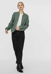 Green bomber jacket with a smooth fabric, front zipper, and ribbed cuffs, paired with black straight-leg jeans and black ankle boots.