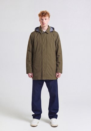 Junger Mann mit lockigem Haar, der einen olivgrünen Kapuzenmantel, dunkelblaue Hose und weiße Sneakers trägt, steht vor einem schlichten hellen Hintergrund.