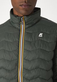 Padded green jacket with a high collar, featuring zigzag stitching, a colorful zip with red, yellow, and blue stripes, and a logo patch.