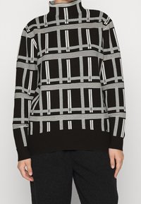 Pull en tricot noir avec motif rayé gris et blanc, col haut et coupe décontractée. Présente des accents de lignes verticales et horizontales.