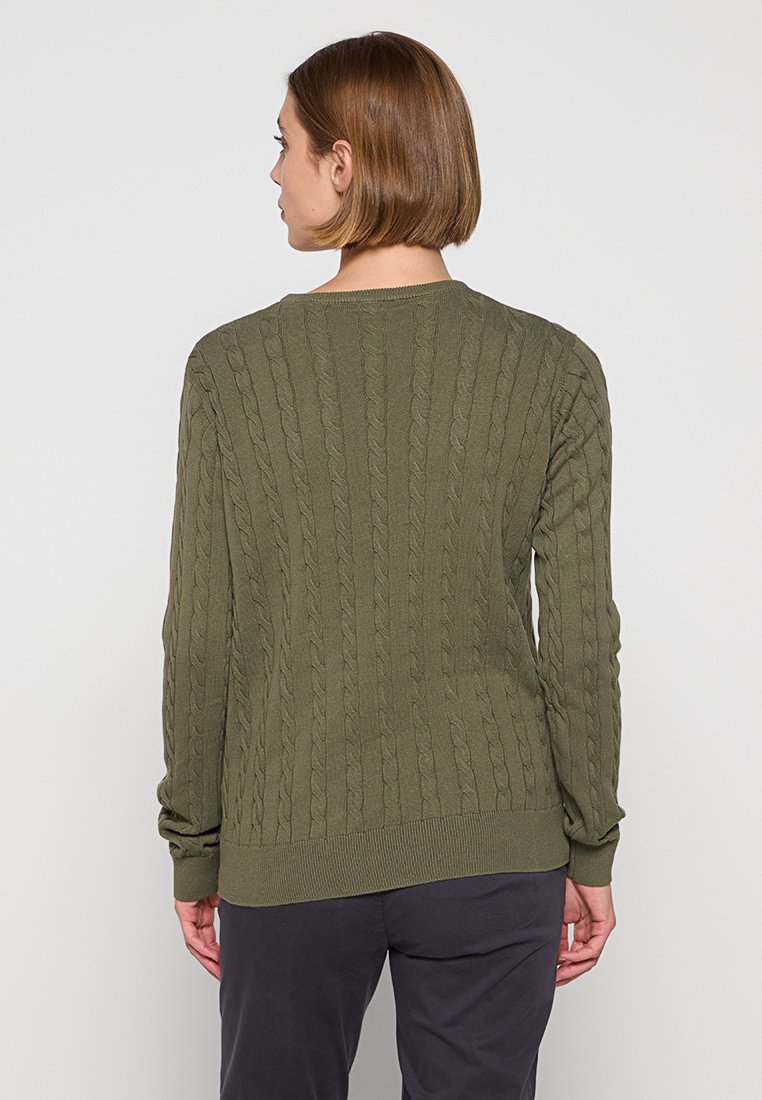 Frau mit kurzen braunen Haaren, die einen olivgrünen Kabelstrickpullover und dunkle Hosen trägt, steht mit dem Rücken zu einer schlichten Hintergrund.