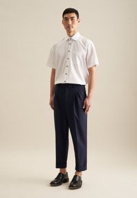 Camicia bianca a maniche corte con tasca sul petto e bottoni neri, abbinata a pantaloni blu a pieghe e scarpe di pelle nere.