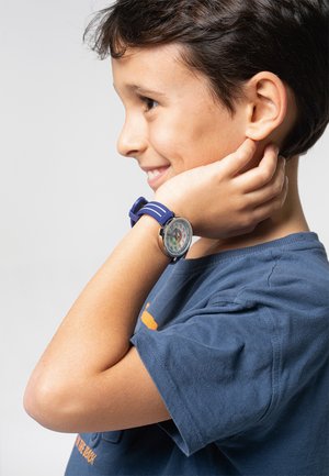 Montre en silicone bleu avec un cadran rond affichant des chiffres et des motifs colorés, portée sur le poignet d'un enfant, accompagnée d'un t-shirt bleu marine.