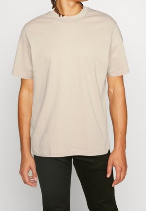 Hombre vistiendo una camiseta de manga corta beige lisa y pantalones negros, de pie contra un fondo blanco.