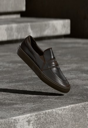 Chaussure à enfiler en cuir brun foncé flottant au-dessus de marches en béton avec une surface texturée et une ombre portée en dessous.