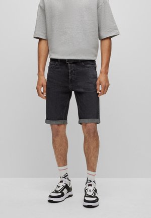 Homme portant un t-shirt gris surdimensionné, un short en jean noir retroussé avec ceinture, des chaussettes blanches et des baskets noir et blanc, debout contre un fond gris.
