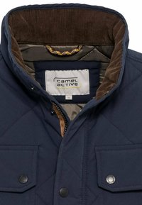 Marineblaues, gestepptes Jacket mit Cordkragen, Reißverschluss, zwei Druckknopftaschen und Stoffetikett im Inneren. Glatte Textur und modernes Design.