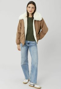 Veste matelassée marron avec un col en fausse fourrure blanche, associée à un pull vert foncé et un jean bleu clair à jambes droites. Baskets blanches avec des accents noirs.