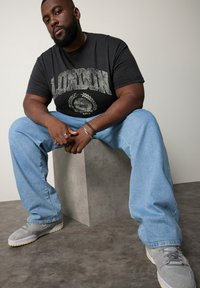 Černé tričko s grafikou "LONDÝN", světle modré džíny a šedé tenisky. Muž sedí na betonovém bloku před neutrálním pozadím.
