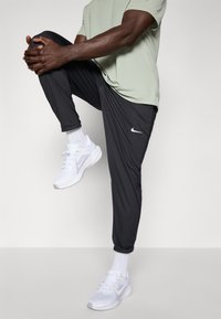 Fekete sportos jogging nadrág fényvisszaverő Nike logóval, fehér sportcipőkkel és világoszöld pólóval párosítva. Simább anyag, karcsúsított fazon.