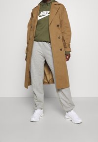 En beige trenchcoat över en grön hoodie med vit Nike-logotyp, grå sweatpants och vita sneakers. Kappan har knappar och en bälte vid midjan.