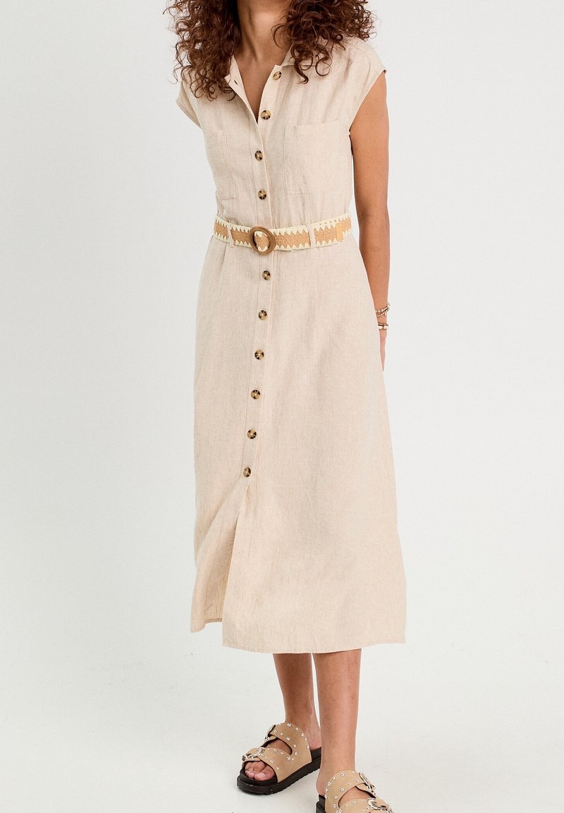 Femme portant une robe beige boutonnée sur le devant, avec une ceinture à motifs et des sandales beiges cloutées, debout devant un fond uni.