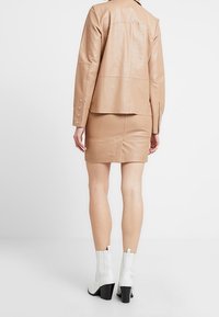 Beige läderjacka med silvernitar och en figurnära beige läderkjol. Modellen bär vita ankelstövlar med en kraftig klack.