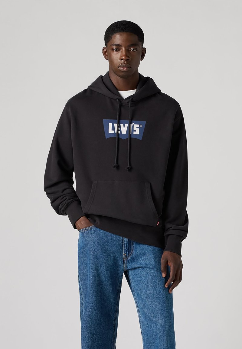 Juodas vilnonis megztinis su mėlynu ir baltu "Levi's" logotipu. Su kengūros kišene, reguliuojamu gobtuvu ir laisvu fasonu. Dėvimas su mėlynomis džinsomis.