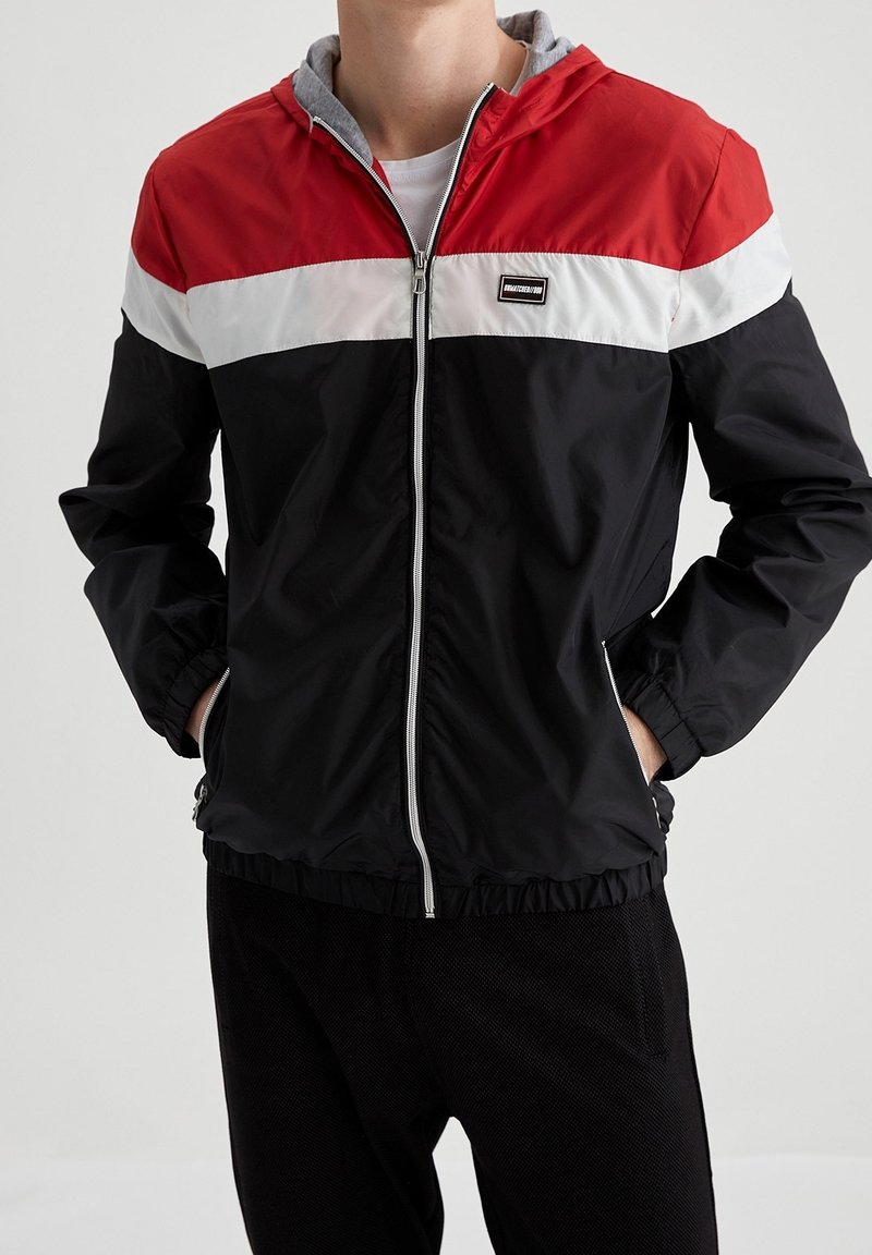 Veste légère noire et rouge avec une fermeture éclair à l'avant, une capuche grise et une bande horizontale blanche. Comprend des poignets et un ourlet élastiques.
