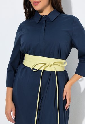 Woman wearing a navy blue button-up dress with three-quarter sleeves and a wide pale yellow belt tied at the waist.