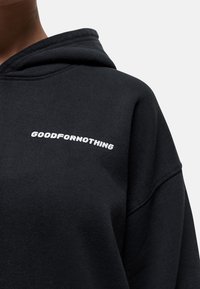 Zwarte hoodie met geborduurde tekst "GOODFORNOTHING" in het wit. De stof lijkt zacht met een ontspannen, oversized pasvorm.