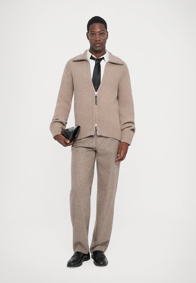 Cardigan bej cu dungi, cu închidere cu fermoar, combinat cu pantaloni gri în carouri și pantofi negri din piele. Ținând o poșetă neagră tip clutch.