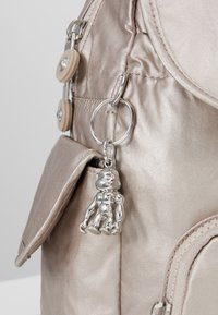 Zaino metallico oro con tessuto testurizzato, dotato di una tasca laterale e di un portachiavi a charm in argento a forma di piccola figura.