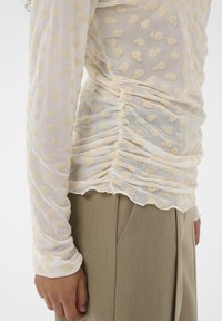 Balts caurspīdīgs tīklojuma tops ar garām piedurknēm, nelieliem dzelteniem ziedu rakstiem un savāktām detaļām sānos, kopā ar bēšām biksēm.