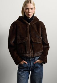 Chaqueta con capucha de pelaje sintético marrón, con dos bolsillos frontales, cierre de cremallera, diseño corto y textura suave, combinada con jeans de mezclilla.