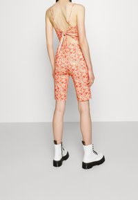 Combinaison florale orange et rose avec un dos ouvert et un détail noué. Comprend des shorts ajustés et est associée à des bottes blanches à lacets.