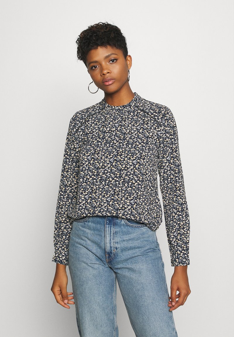 Blusa de manga longa floral em azul-marinho com pequenos padrões em creme e amarelo, decote redondo e tecido leve, combinada com jeans de cintura alta.