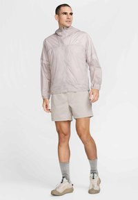 Lätt, ljuslila zip-jacka med huva, featuring elastiska muddar och en strömlinjeformad design, bärs över beige shorts och grå strumpor.