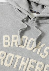 Felpa con cappuccio grigia caratterizzata da ricamo testurizzato color crema con la scritta "BROOKS BROTHERS". Il tessuto presenta una consistenza morbida, simile al fleece.