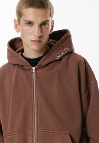 Hnedá mikina s dlhým zipsom a veľkou kapucňou, vyrobená z mäkkého materiálu. Má minimalistický dizajn s vyšitým vzorom na golieri.