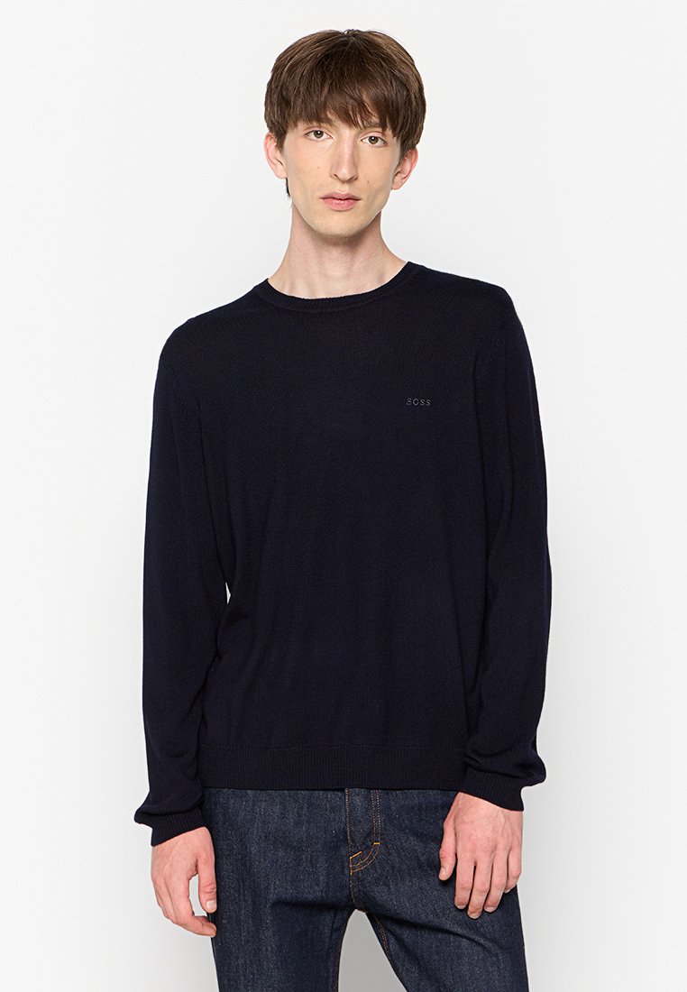 Jongeman met kort bruin haar, draagt een zwart sweater met lange mouwen en donkere spijkerbroek, staande tegen een witte achtergrond.