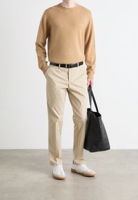 Uomo che indossa un maglione cammello, pantaloni beige, sneakers bianche, mentre tiene una borsa a tracolla nera in pelle, in piedi su un pavimento grigio contro un muro bianco.