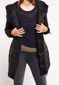 Sort pufferjakke med quiltet design, lynlåslukning og stor hætte. Båret over en navy striktrøje og en beige top, parret med mørke jeans.