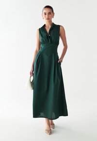 Grünes ärmelloses Kleid mit kragenförmigem Ausschnitt, taillierter Taille, A-Linie Silhouette, seitlichen Taschen und strukturiertem Stoff. Getragen mit beigen Schuhen.