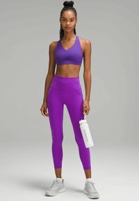 Un reggiseno sportivo viola e leggings a vita alta realizzati in materiale elastico. La modella tiene in mano una bottiglia d'acqua bianca etichettata "lululemon", indossando scarpe da ginnastica grigie.