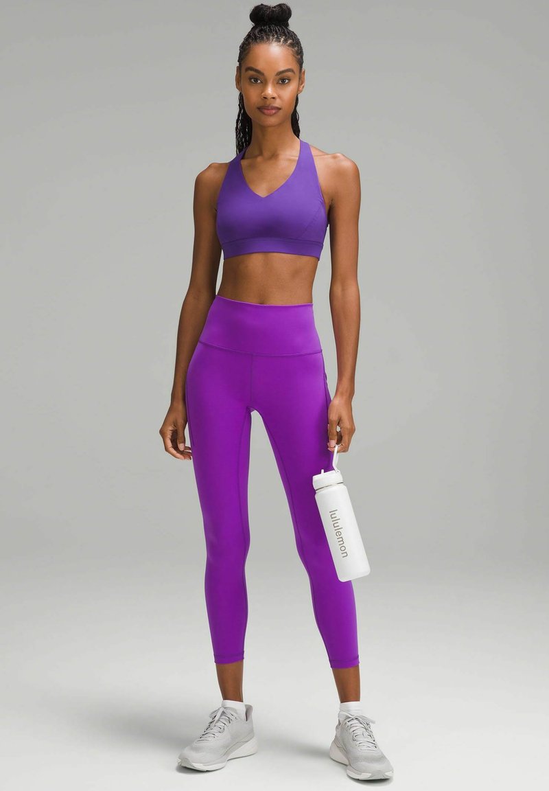 Un reggiseno sportivo viola e leggings a vita alta realizzati in materiale elastico. La modella tiene in mano una bottiglia d'acqua bianca etichettata "lululemon", indossando scarpe da ginnastica grigie.