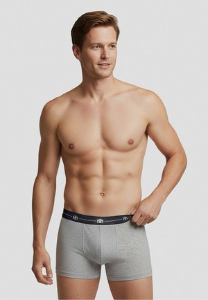 Hemdloser Mann mit heller Haut und braunen Haaren, trägt graue Boxershorts, richtet den Bund, steht vor schlichtem hellem Hintergrund.