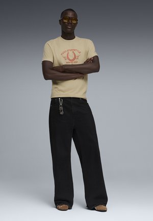 Mężczyzna ubrany w beżowy t-shirt z nadrukiem, oversize’owe czarne spodnie z przypiętymi kluczami, brązowe buty oraz okulary przeciwsłoneczne o bursztynowym zabarwieniu, stoi z założonymi skrzyżowanymi ramionami.